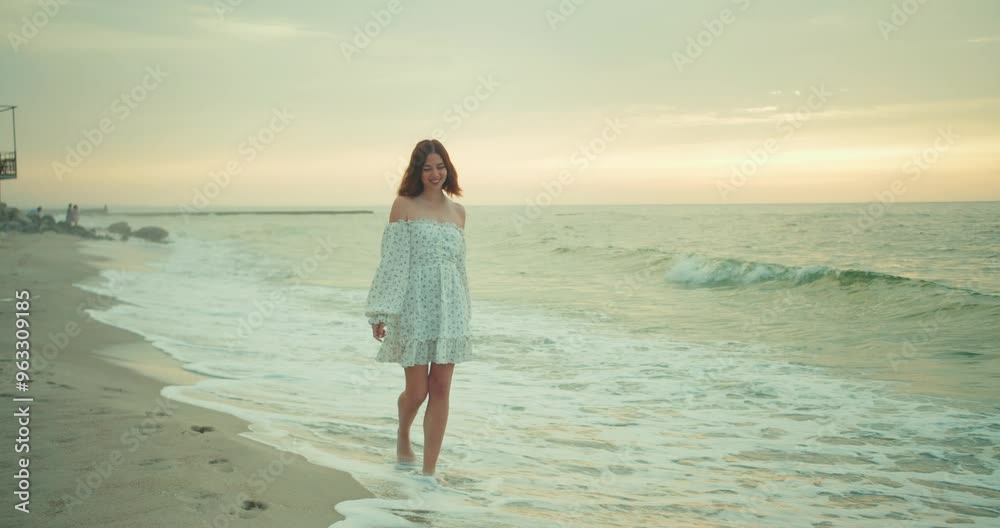 A happy and confident brunette girl in a white dress walks along the water along the sea on the beach early in the morning A happy and confident brunette girl in a white dress walks along the water along the sea on the beach early in the morning
