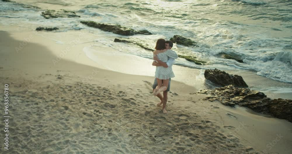 Top view of a happy couple, brunette guy lifts his girlfriend in a white dress and spins her while dancing together on the beach in the morning near the sea Top view of a happy couple, brunette guy lifts his girlfriend in a white dress and spins her while dancing together on the beach in the morning near the sea