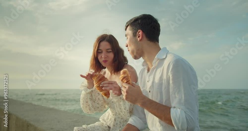 Happy brunette girl in a white dress eats a croissant with her boyfriend during her date on the beach in the morning