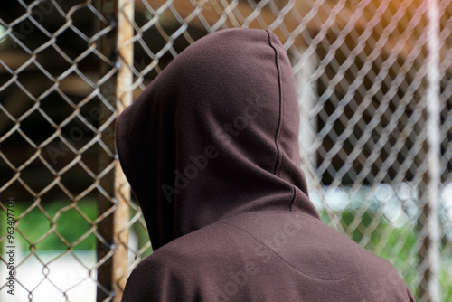 male teenager wearing a brown hoodie stood facing a steel mesh fence. Both hands cling to the fence, showing the back.