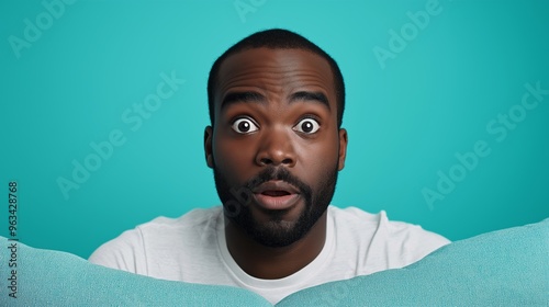 A man with a surprised expression on his face is looking at a pillow. The pillow is blue and has a pattern of lines on it. The man is standing in front of the pillow