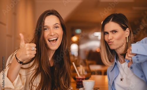 Fotografie Two women sisters friends rest in cafe, she show like and dislike, thumb up and thumb down, disagreement and agreement expression