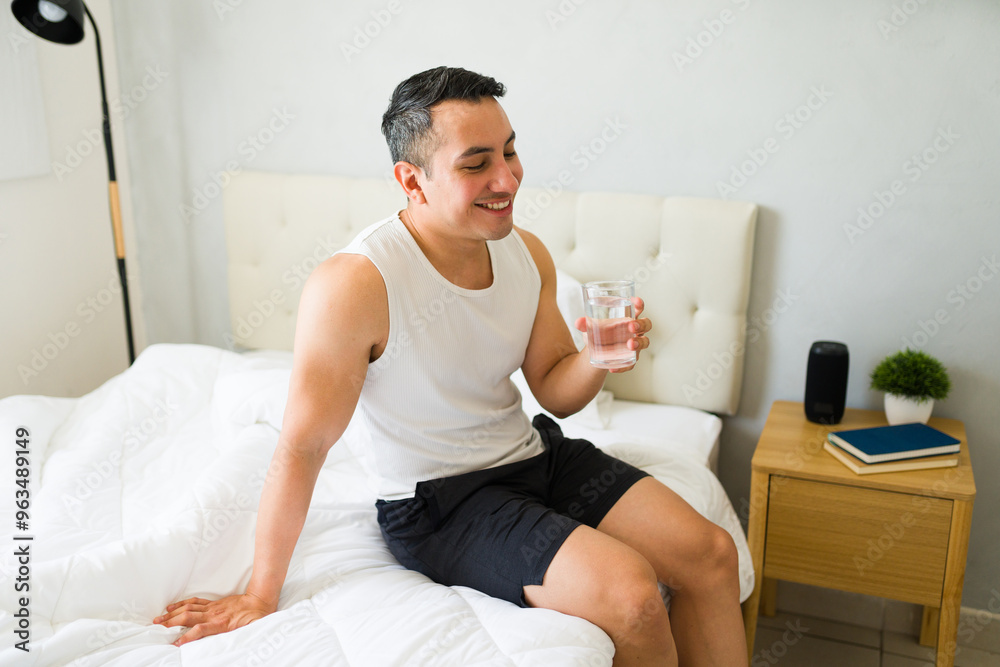 Smiling hispanic man sitting on bed while drinking a glass of fresh water after waking up in the morning Smiling hispanic man sitting on bed while drinking a glass of fresh water after waking up in the morning