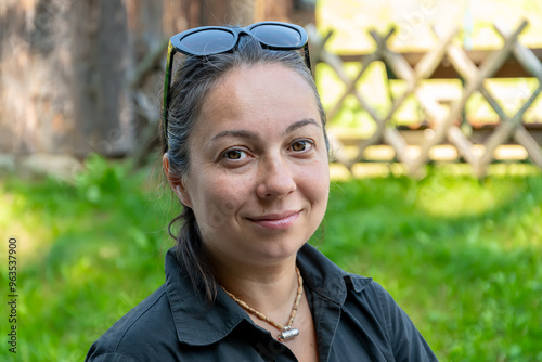 A 40-45 year old woman wearing sunglasses on her head smiles and poses in a serene green outdoor area surrounded by trees and a wooden fence in the midday light. photo