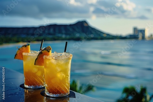 Two drinks with pineapple slices in them are on a table by the ocean. The drinks are in tall glasses and the ocean is in the background photo