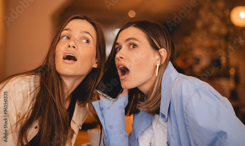 Shocked beautiful young woman overhearing conversation of cheerful female with mobile phone sitting in cafe. Two women sisters or friends listen phone call, look shocked, wow. photo