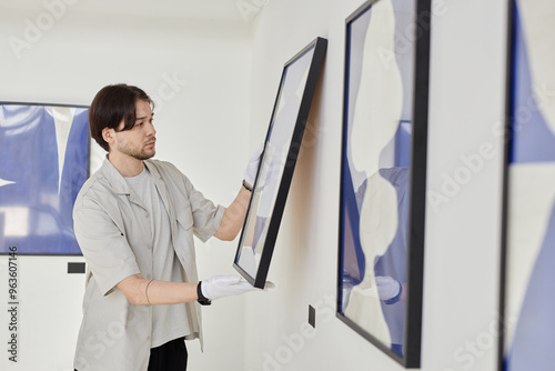 Minimal side view portrait of young Asian man working as art curator and hanging paintings on wall while planning modern art gallery exhibition copy space