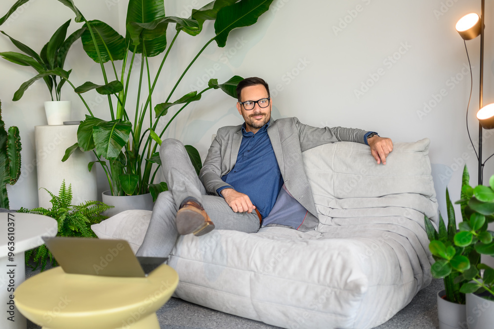 Thoughtful male CEO in eyeglasses looking away while sitting comfortably on sofa at modern workplace Thoughtful male CEO in eyeglasses looking away while sitting comfortably on sofa at modern workplace