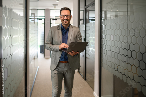 Confident male professional working over laptop and smiling at camera while standing in office corridor photo