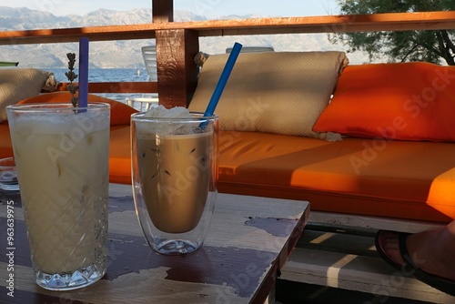 Glasses with non alcoholic Pina Colada and ice coffee with blue straws placed on decorative table in beach bar, orange comfy sofa, bay and shore with shore mountain line in background. Afternoon sun photo