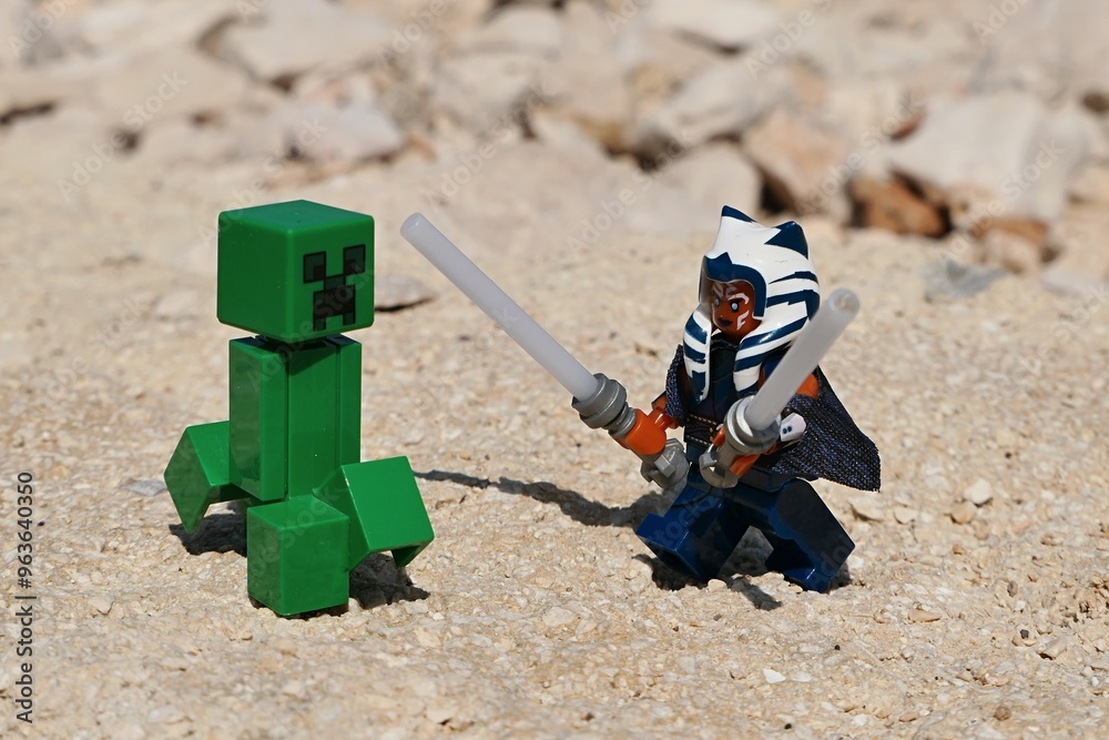Naklejka premium LEGO Star Wars figure of female Togruta Jedi Ahsoka Tano with two lightsabers ready to fight with green explosive LEGO Minecraft mob Creeper on shoreline rocks, summer daylight sunshine. 