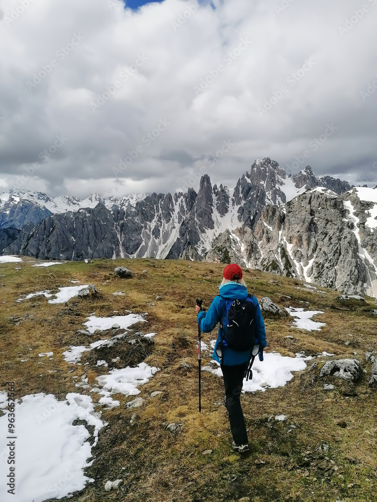 A lone hiker wearing a blue jacket, red hat, and carrying trekking poles traverses a grassy area among the snowy peaks of the Dolomites on a cloudy spring day. A lone hiker wearing a blue jacket, red hat, and carrying trekking poles traverses a grassy area among the snowy peaks of the Dolomites on a cloudy spring day.