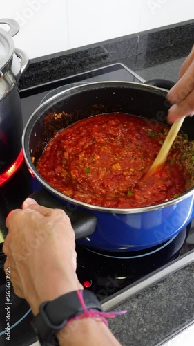 Vertical shot of hands of woman cooking bolognese sauce, mixing fried tomato, minced meat, zucchini, slices of mushrooms and diced onion with a wooden spoon, next to stainless saucepan on an electric 