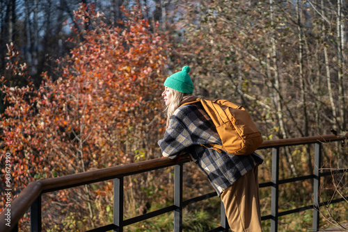 Tranquil woman with backpack leaning on fence in autumn park, soaking up sunlight. Female explorer with closed eyes resting, solo travel at relaxed pace, contemplative living, slow life, time alone photo