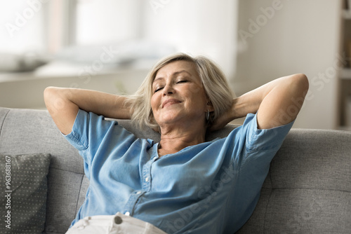 Fotografie Calm older woman leaning back on sofa with hands behind head, resting with eyes