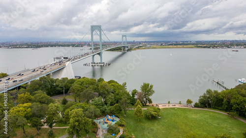 A scenic view of the Whitestone Bridge crossing the East River, connecting urban and natural landscapes. The nearby park offers green spaces with river views, with the horizon and the bustling city of