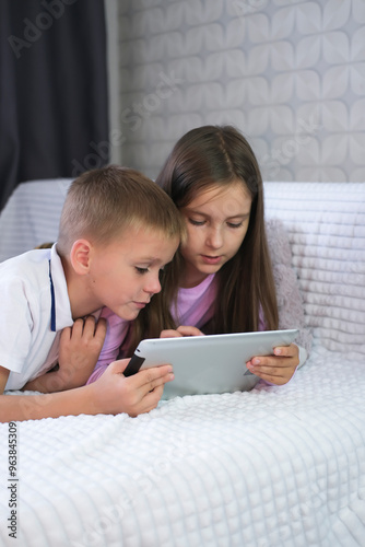 Brother and sister are lying on sofa at home and playing game on digital tablet. Children watch videos online on digital tablet. Internet. Vertical.