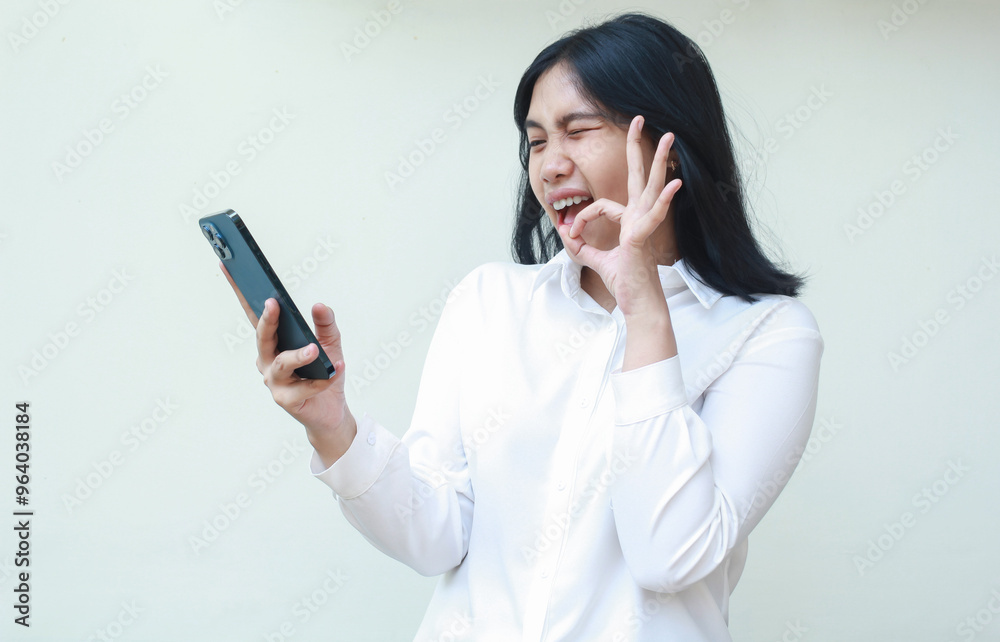 delighted asian young woman secretary holding mobile phone showing ok sign, okay symbol, wearing white formal suit shirt, flirting blink eye to screen in video call, agree gesture delighted asian young woman secretary holding mobile phone showing ok sign, okay symbol, wearing white formal suit shirt, flirting blink eye to screen in video call, agree gesture
