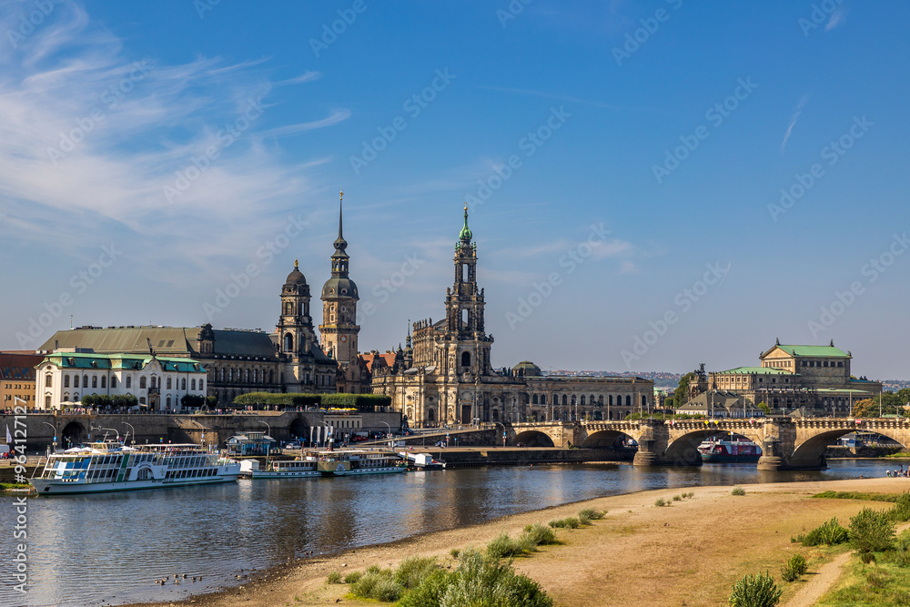 Fototapeta premium Blick auf die Dresdner Altstadt mit der Katholischen Hofkirche (auch bekannt als Kathedrale St. Trinitatis) im Zentrum. Im Vordergrund fließt die Elbe, auf der mehrere Schiffe zu sehen sind. 