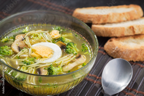 Noodle soup with broccoli, mushrooms, leek and hard-boiled egg in a glass bowl.