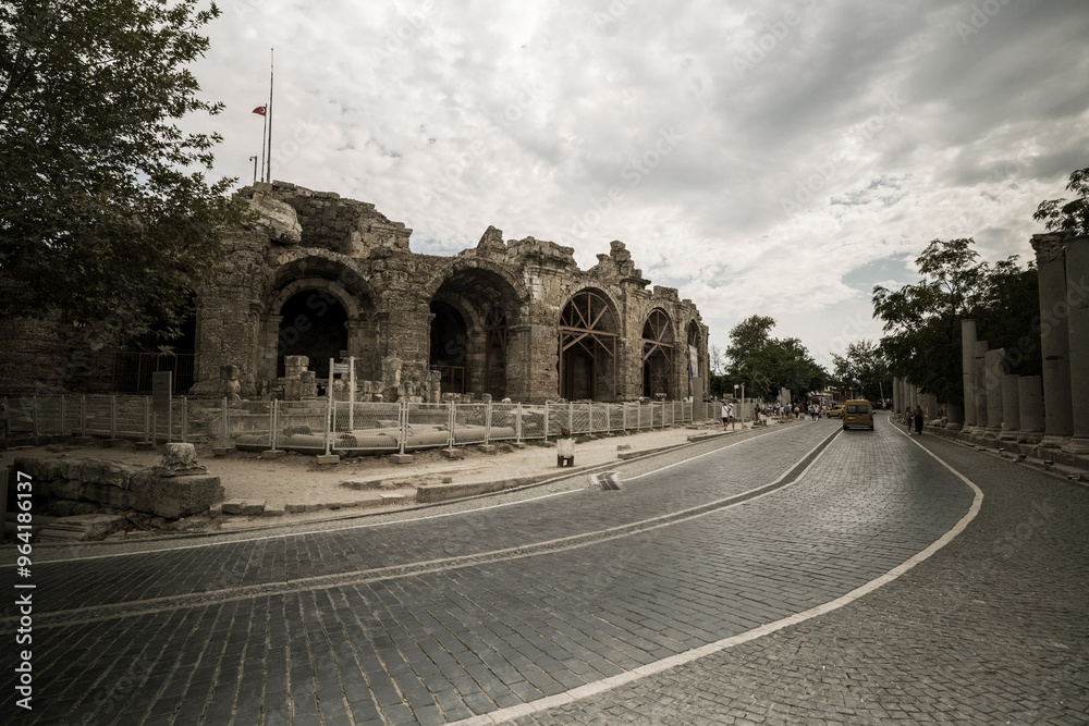 A cobblestone road curves past the ancient amphitheater in Side, Turkey. The amphitheater's weathered stone facade stands tall against a cloudy sky.