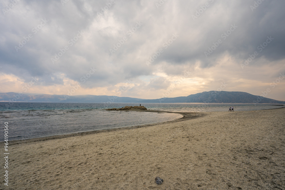 A tranquil scene at Lake Salda, Turkey, featuring a sandy beach curving into the distance, clear waters, and mountains under a cloudy sky.