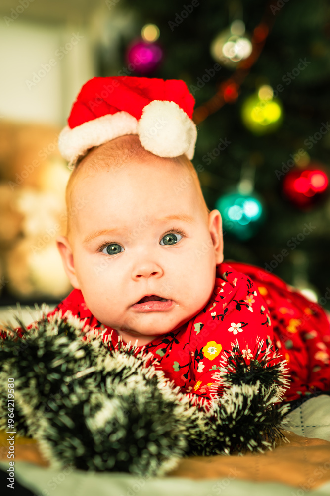 Newborn baby boy in a red Santa hat lies on the bed near the Christmas tree Newborn baby boy in a red Santa hat lies on the bed near the Christmas tree