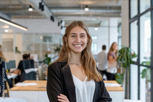 A smiling young professional standing confidently in a modern office, with colleagues working in the background, embodying success and collaboration. Free Download A smiling young professional standing confidently in a modern office, with colleagues working in the background, embodying success and collaboration. - GFXLoad