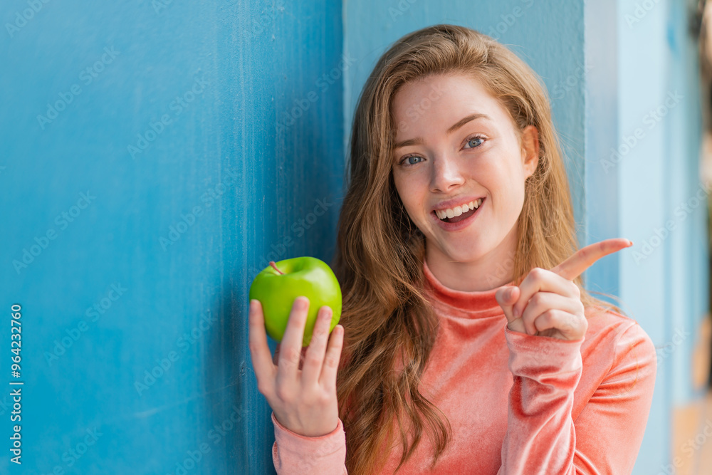 Young redhead woman with an apple at outdoors surprised and pointing finger to the side Young redhead woman with an apple at outdoors surprised and pointing finger to the side
