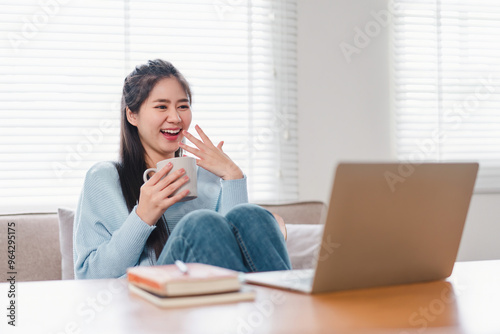 Smiling Asian woman typing on laptop while sitting at home, focused on work or study in a comfortable setting.