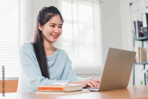 Smiling Asian woman typing on laptop while sitting at home, focused on work or study in a comfortable setting.