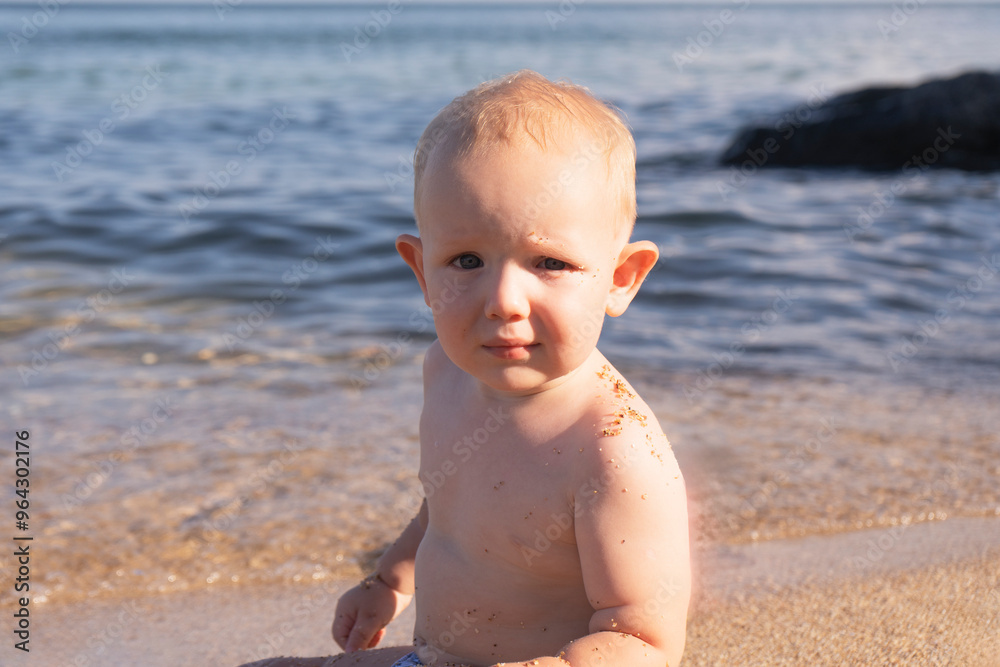 Portrait of a cute eared little boy against the backdrop of the sea on a bright sunny day. Cute baby playing with sand on the seashore. Close up. Portrait of a cute eared little boy against the backdrop of the sea on a bright sunny day. Cute baby playing with sand on the seashore. Close up.