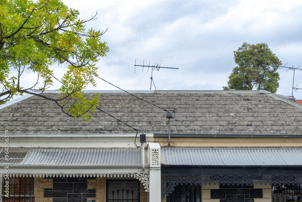 Close-up view of two adjacent Victorian-era suburban houses with roofs covered in weathered slate tiles, cast iron laceworks, and a television antenna on top in Australia. Close-up view of two adjacent Victorian-era suburban houses with roofs covered in weathered slate tiles, cast iron laceworks, and a television antenna on top in Australia.