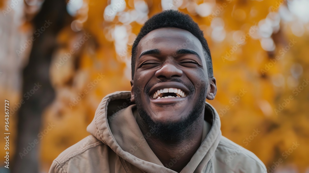 A close-up of a man laughing with his eyes closed, against a blurred background of autumn leaves.
