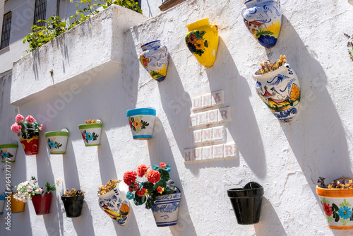 A white wall decorated with colorful ceramic flower pots in vibrant designs. Red flowers and a sign reading "UNA FLOR UNA MUJER" as the focal point. Olvera, Andalusia, Spain