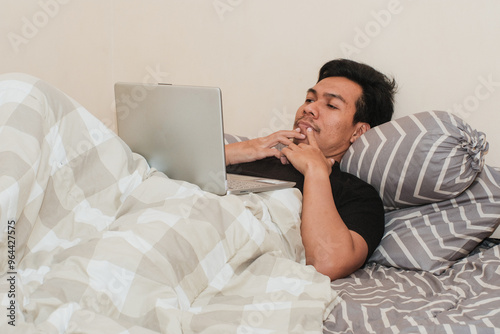 A Southeast Asian man in bedroom and laptop for work from home, serious or thingking in bed for freelance idea. Technology, entrepreneur and remote worker for online job. photo