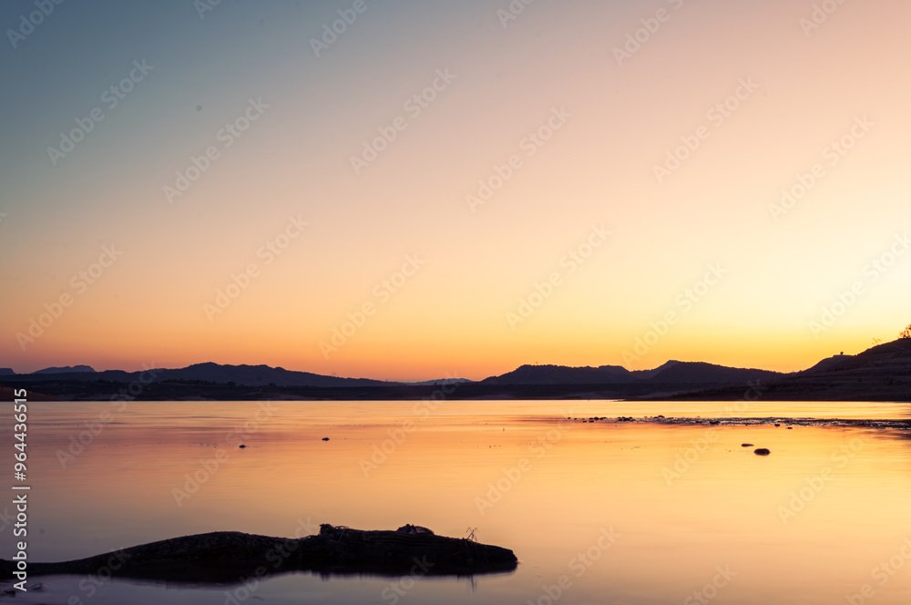 Tranquil lake with still water reflects the golden light of setting sun against backdrop of distant mountains Tranquil lake with still water reflects the golden light of setting sun against backdrop of distant mountains