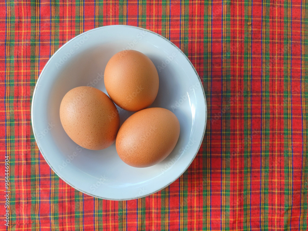 Three brown-colored boiled chicken eggs nestled in a small white bowl, set against the vibrant backdrop of a red and green plaid tablecloth, creating a cozy and inviting presentation Three brown-colored boiled chicken eggs nestled in a small white bowl, set against the vibrant backdrop of a red and green plaid tablecloth, creating a cozy and inviting presentation