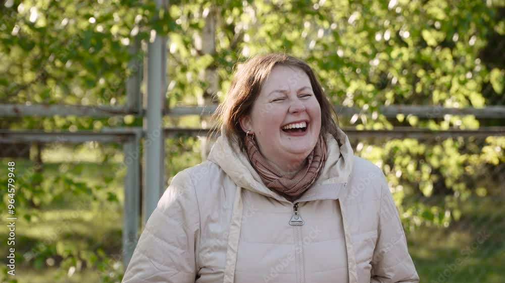 Portrait of middle-aged nordic woman laughing heartily during conversation outdoors. Curvy lady in jacket laughs loudly, cheerfully in sunny park, handheld shot. Portrait of middle-aged nordic woman laughing heartily during conversation outdoors. Curvy lady in jacket laughs loudly, cheerfully in sunny park, handheld shot.