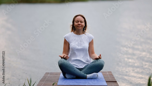 Young Caucasian woman sitting in lotus position on bridge by lake during rain. Woman enjoying fresh air. Outdoor meditation photo