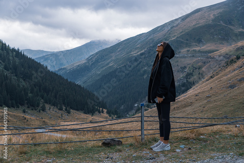 A woman in a hoodie stands on a mountain path, looking up at the cloudy sky. The rugged Carpathian landscape and pine-covered slopes stretch out behind her, creating a peaceful and contemplative scene photo