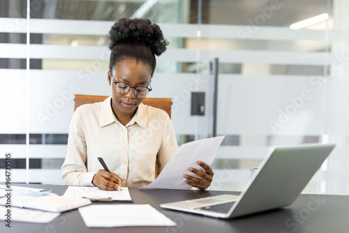 Professional African American businesswoman working diligently at desk with laptop and documents, showcasing dedication and focus. Image reflects modern office setting