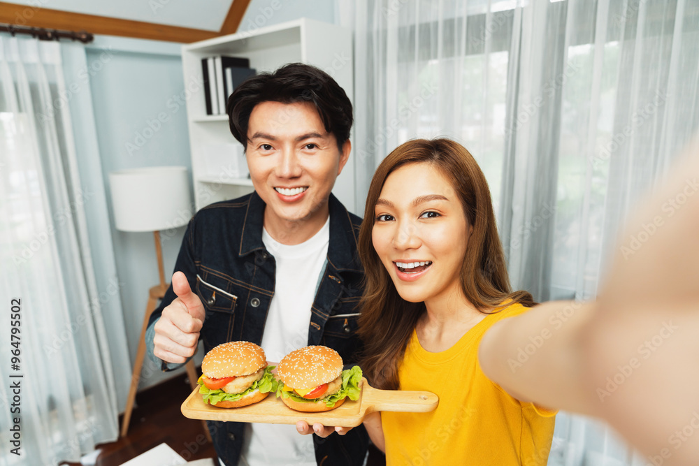 Smiling host chef influencers taking photo with smartphone by selfie straight view holding double hamburger on cooking show special easy dish homemade on social media channel live streaming. Infobahn.