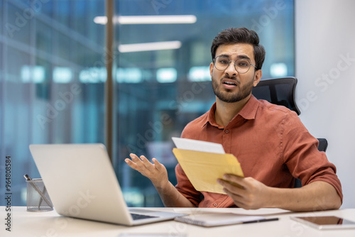 Office worker looks distressed after opening unexpected mail. Seated at desk with laptop and phone, expression reflects surprise and concern. Scene captures challenging moment in professional setting