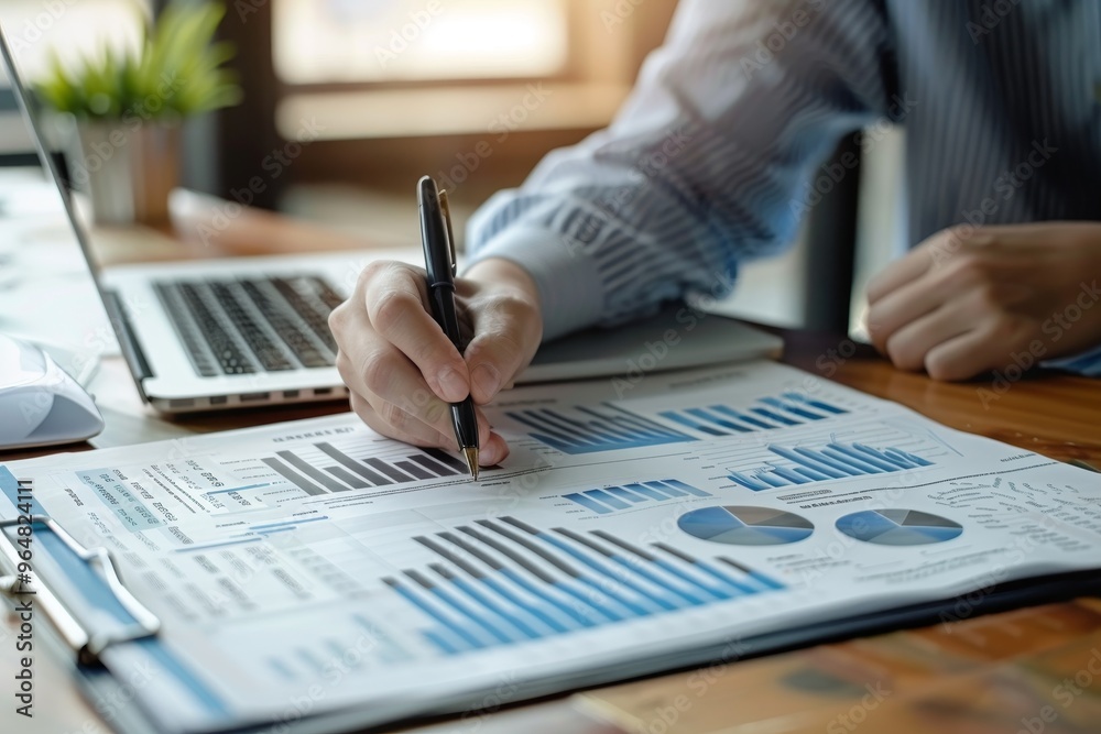 A business professional takes notes on detailed charts and graphs while working on a laptop in a bright and organized office setting during the morning. A business professional takes notes on detailed charts and graphs while working on a laptop in a bright and organized office setting during the morning.