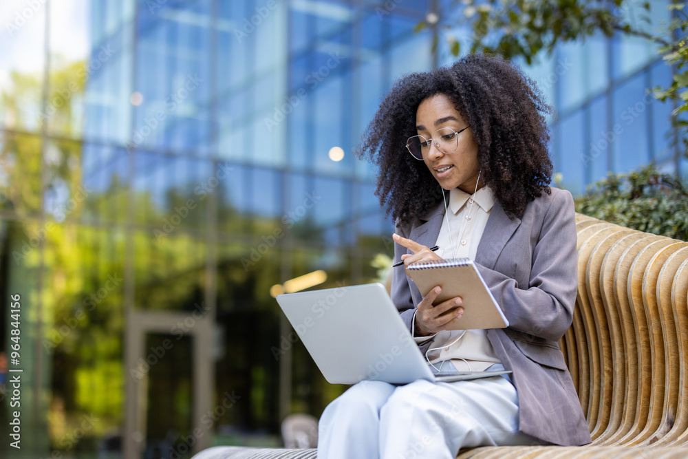 Fototapeta premium African American businesswoman participates in an online video course outside. She engages with instructor via laptop, takes notes in notebook uses for video call, enhancing professional skills
