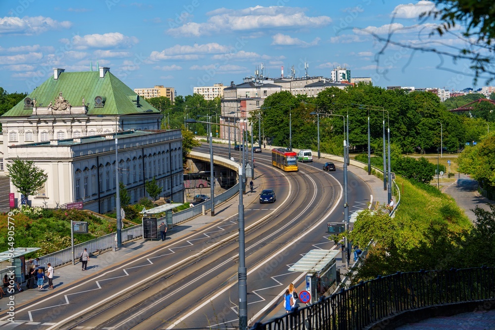 This image depicts a wide street in Warsaw, Poland, featuring a tram near a historic building, trees lining the street, cars present, and a blue sky with clouds above tall buildings. This image depicts a wide street in Warsaw, Poland, featuring a tram near a historic building, trees lining the street, cars present, and a blue sky with clouds above tall buildings.