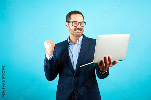 Studio portrait of a mature man standing in smart outfit holding a laptop making success gesture 