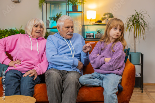 Angry grandfather and grandmother scolding granddaughter for bad behavior and disobedience on sofa, raising voice, scream at little grandchild. Difficulties of upbringing, misbehaved child concept photo