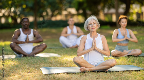 Fotografi Relaxed, immersed in meditation senior woman sitting on exercise mat in Padmasana position with hands clasped in namaste gesture during group yoga class in serene sunlit summer park